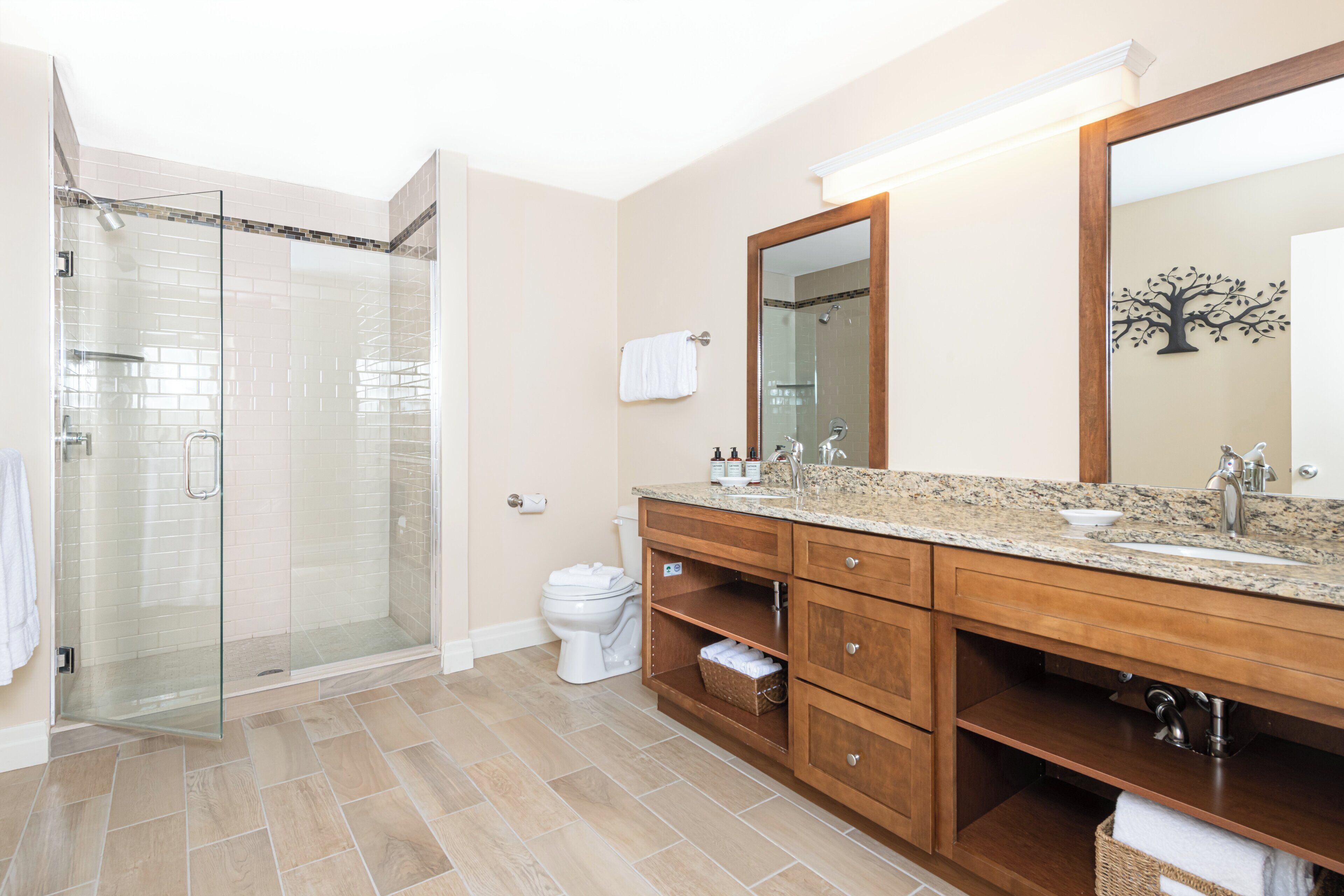 Un baño moderno con cabina de ducha de vidrio, tocador de madera con un espejo grande y suelo de baldosas color beige.