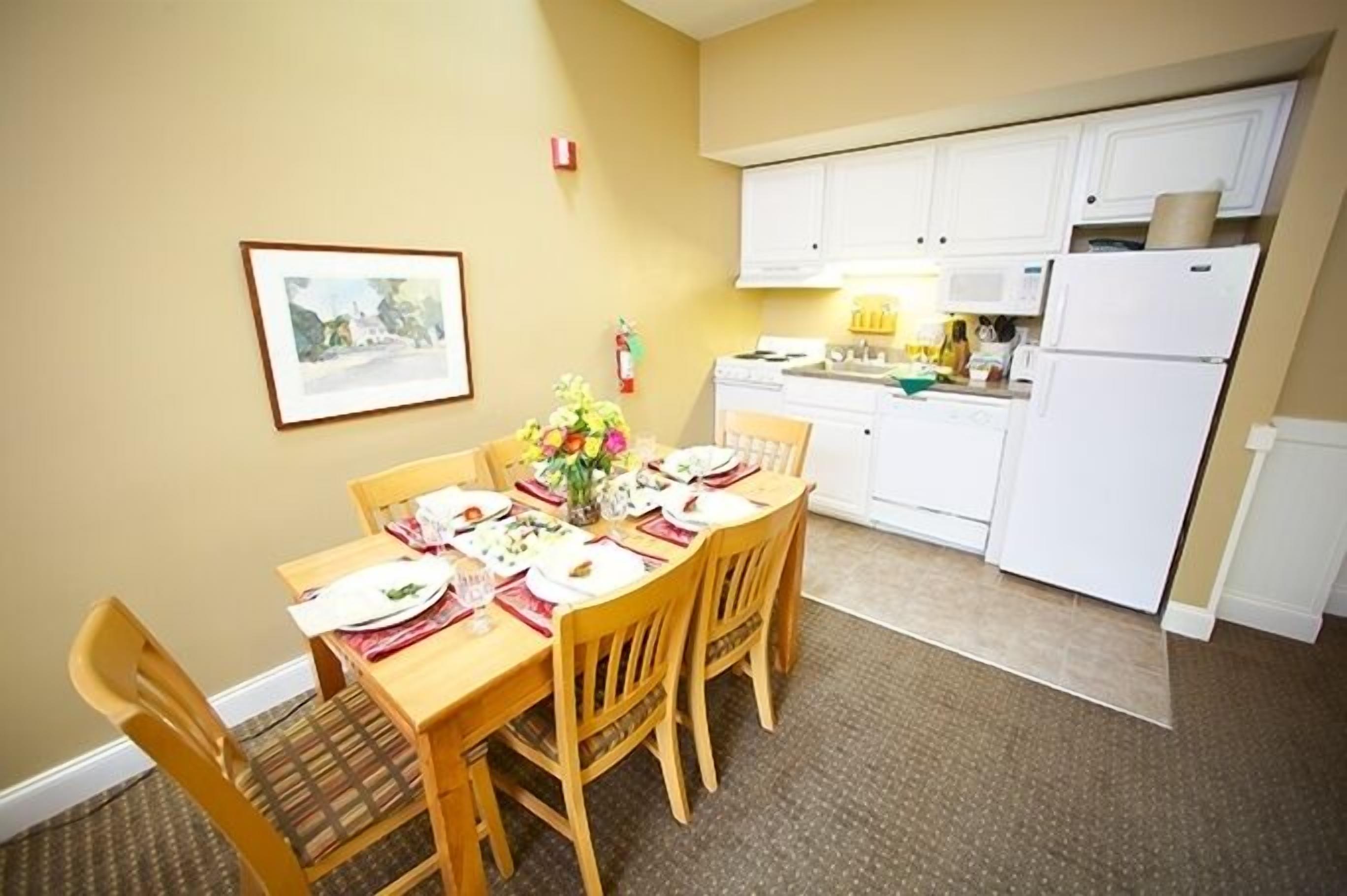 A compact kitchen with white cabinets, a microwave, and a refrigerator. A dining table with chairs is set up in the foreground.