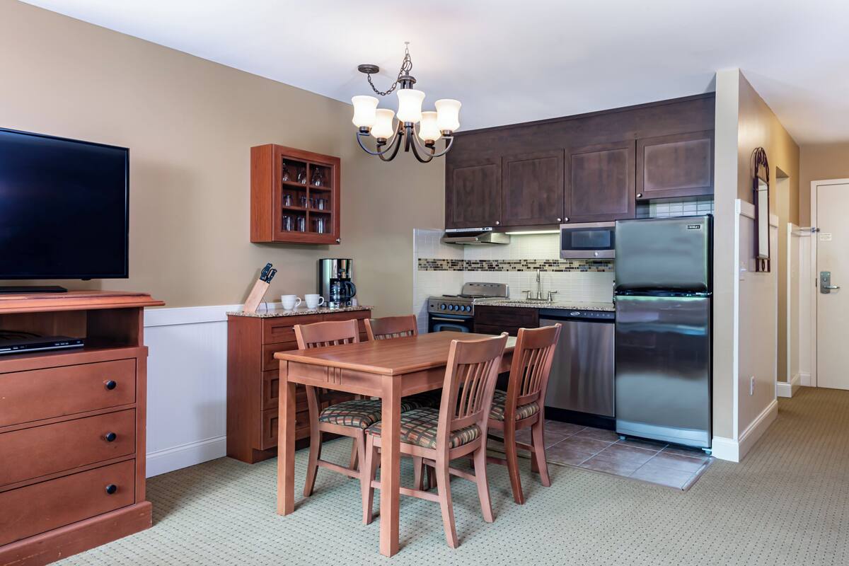 A compact kitchen with wooden cabinets, a stainless steel refrigerator, a microwave, and a coffee maker. There is a dining table with chairs and a television on a stand.