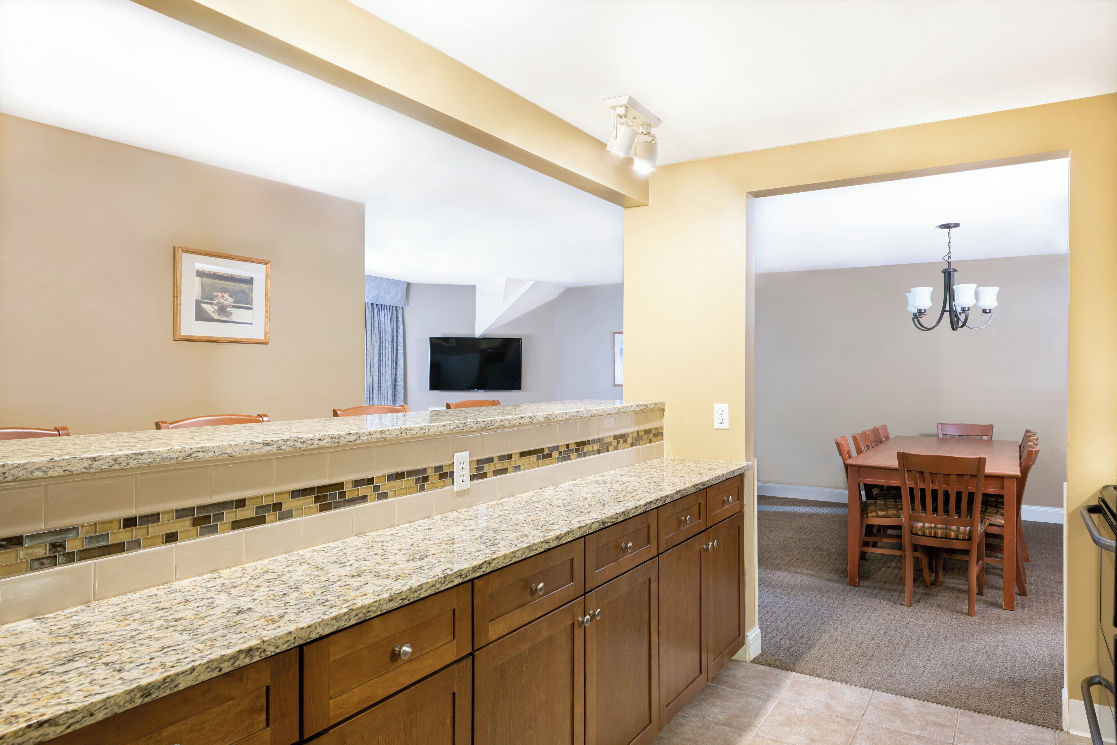A kitchen with granite countertops, wooden cabinets, a dining area with a table and chairs, and a TV mounted on the wall.