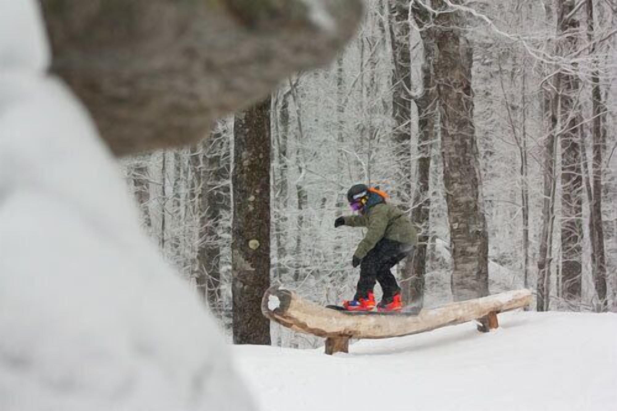 A person snowboarding on a wooden rail in a snowy forest.