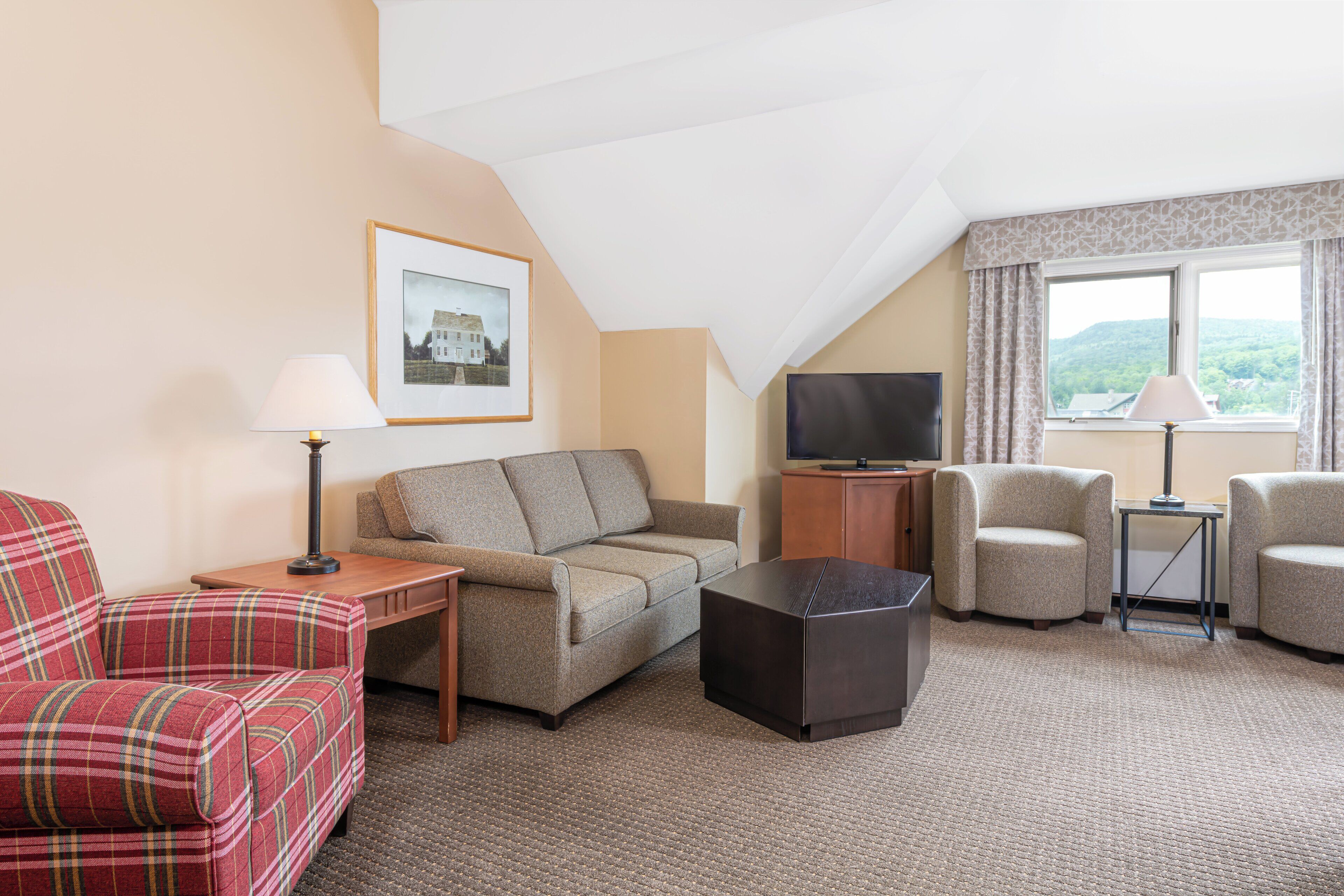 A hotel room with a sofa, armchair, a small table, a TV, and a window with curtains.