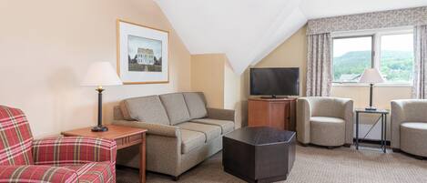 A hotel room with a sofa, armchair, a small table, a TV, and a window with curtains.