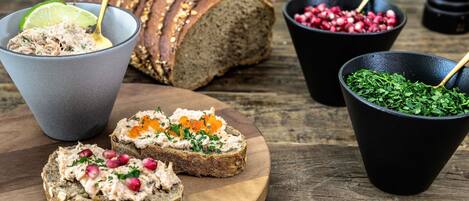 A wooden board with slices of bread topped with a creamy spread, a bowl of pomegranate seeds, and a bowl of chopped herbs.