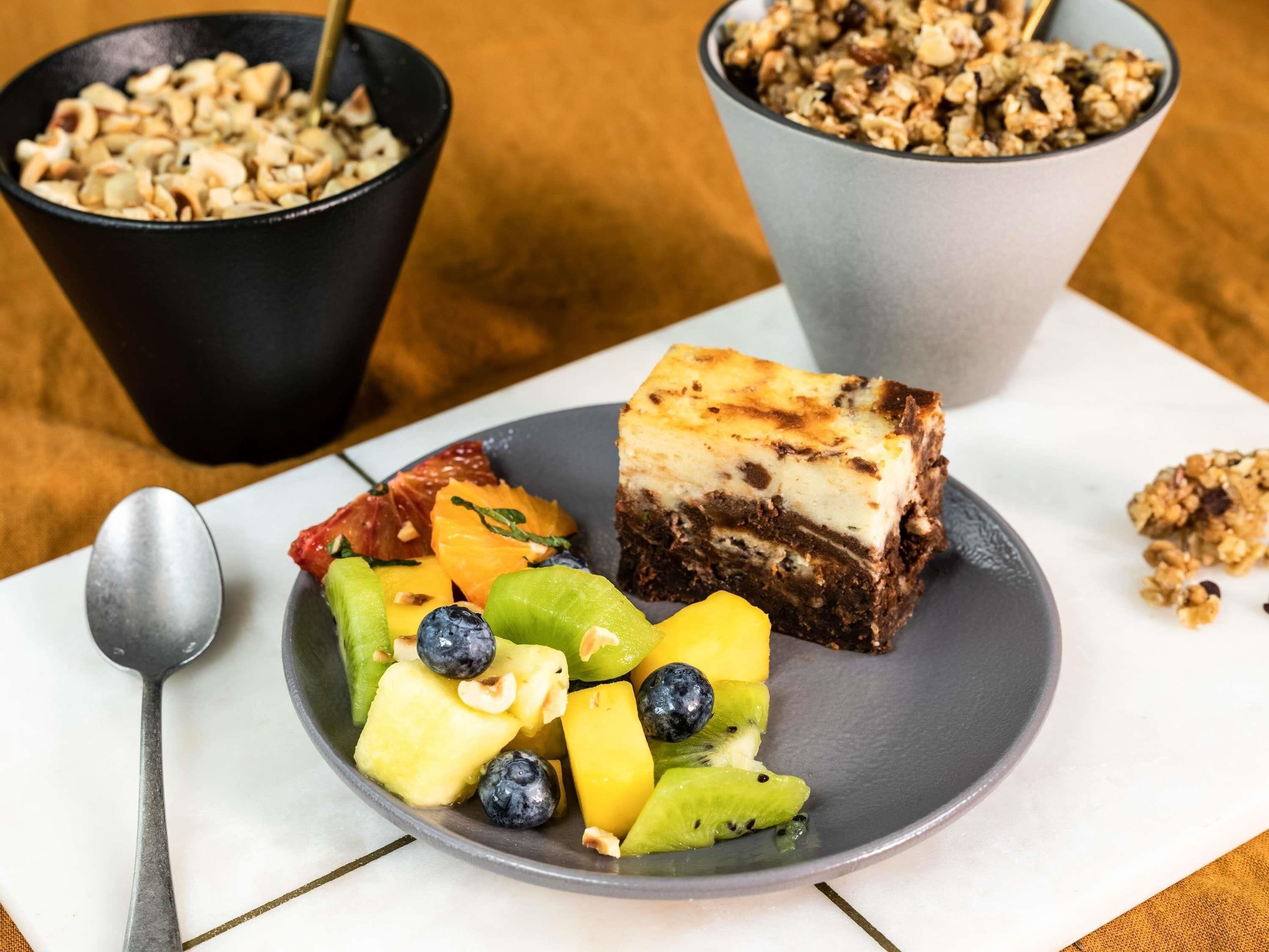 A plate with a slice of cheesecake and assorted fruit, a bowl of granola, and a spoon on a table.