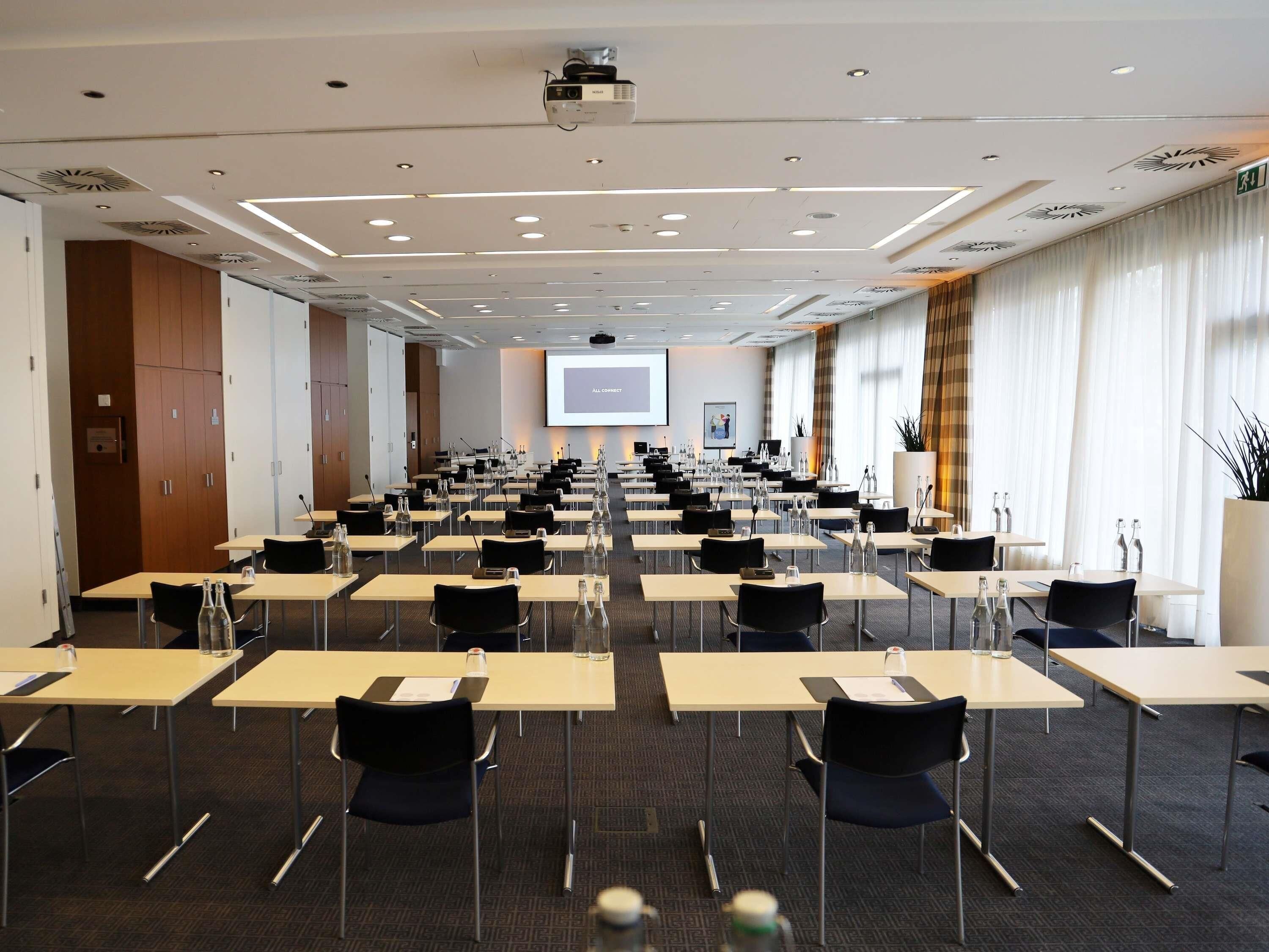 A conference room with rows of tables and chairs, a projector screen, and a large window with curtains.
