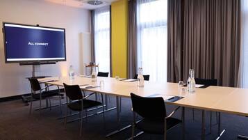 Ett konferensrum med ett långt bord, stolar, en projektorduk och ett fönster med gardiner.