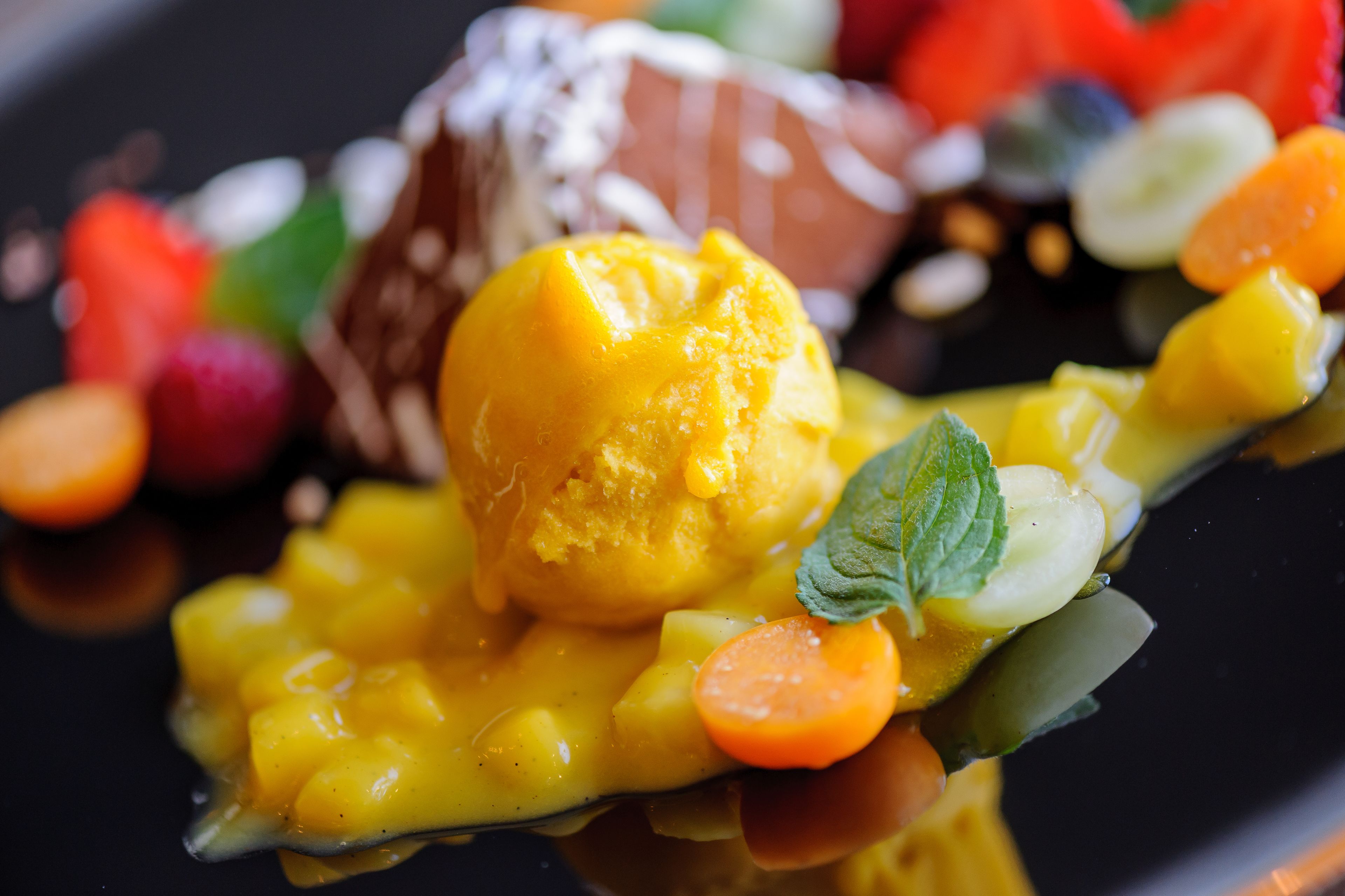 Postre con puré de mango, hojas de menta y una variedad de frutas sobre un plato negro.