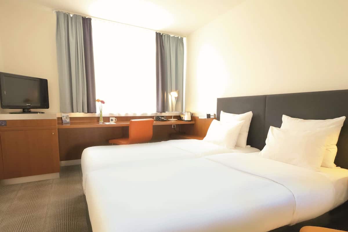 A hotel room with a large bed, a desk, a chair, a television, and a window with curtains.