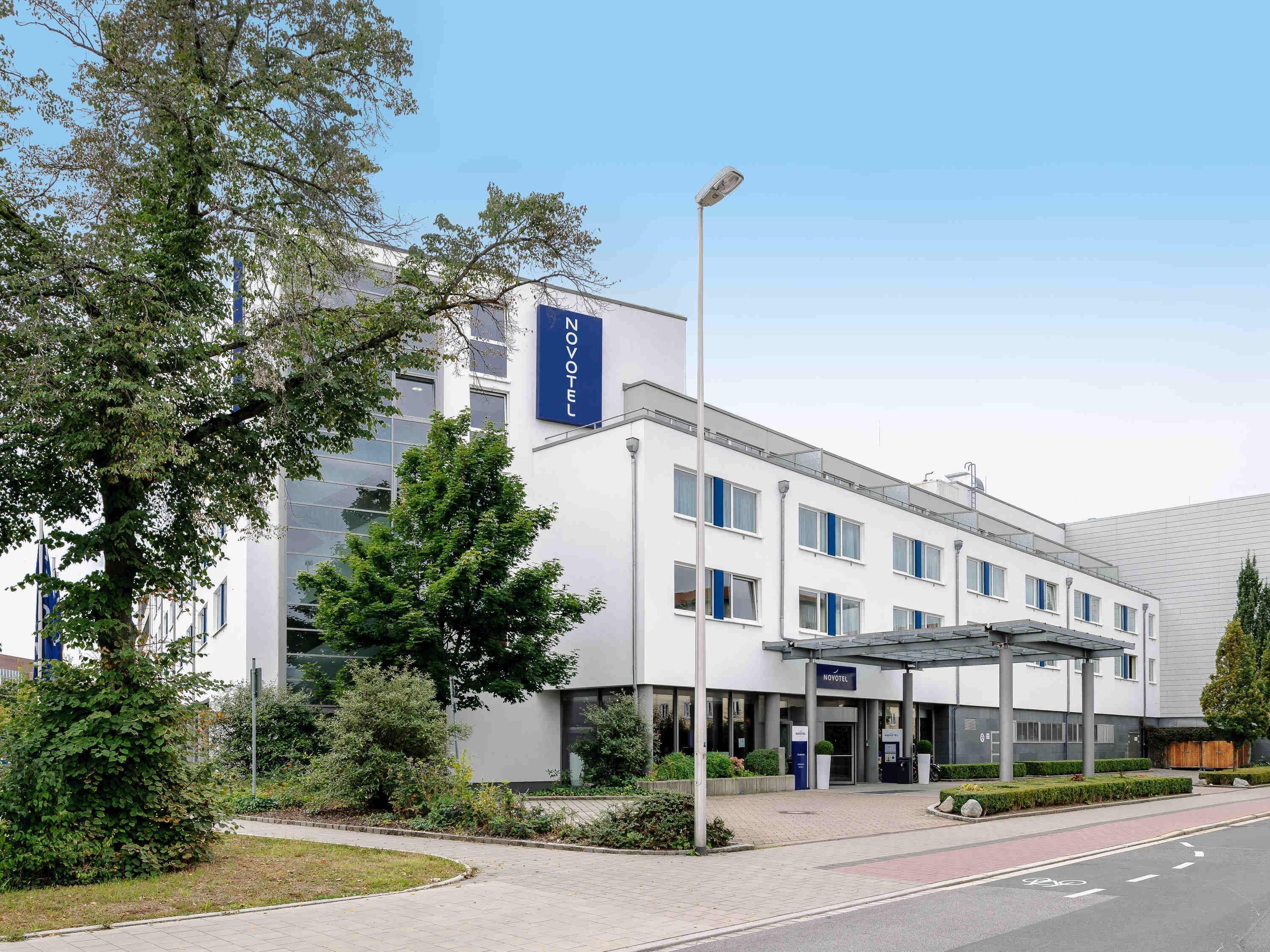 Een modern hotel met een opvallend 'NOVOTEL'-bord, een overdekte ingang en een aangelegde tuin met bomen en struiken.