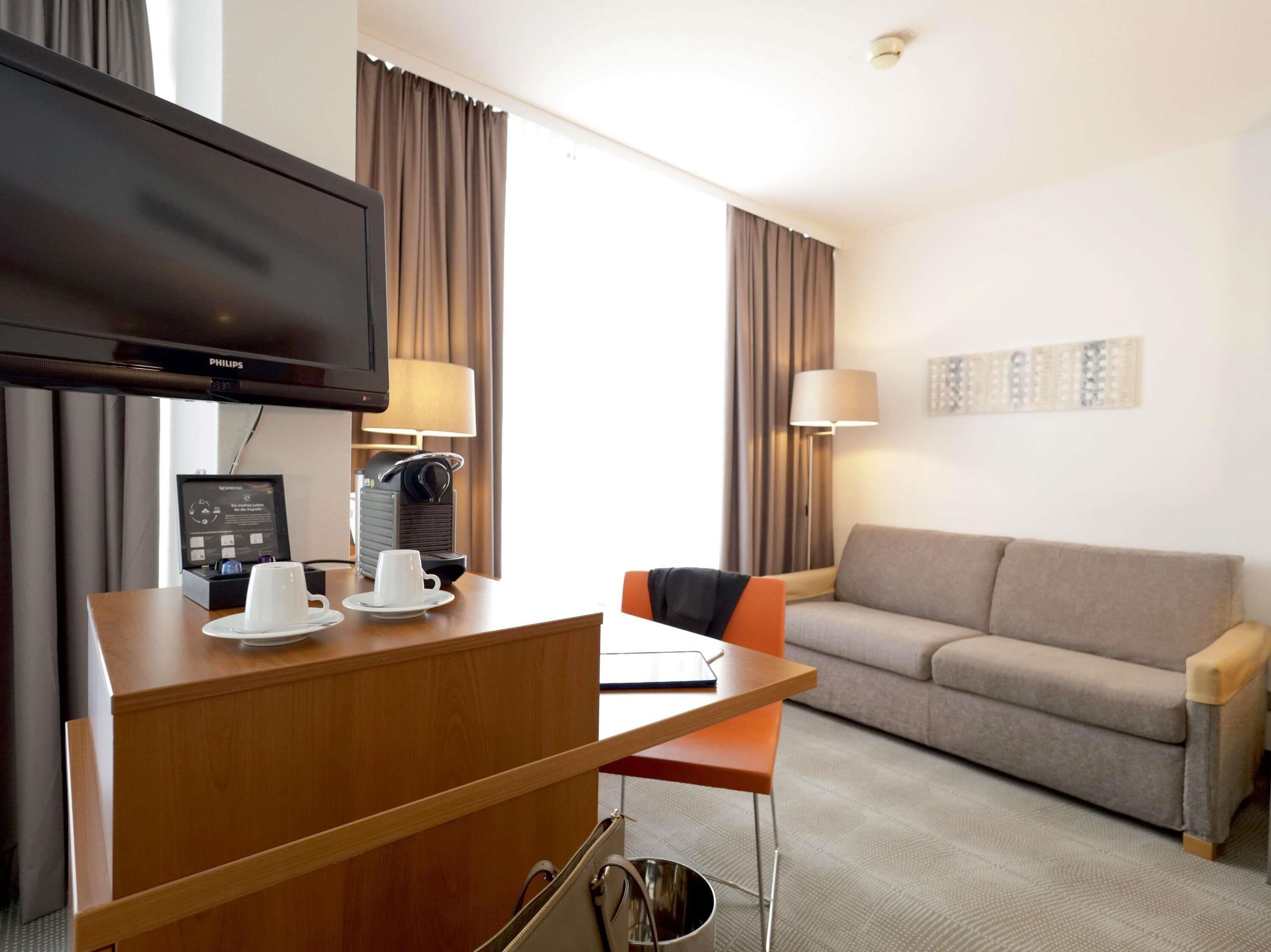 A hotel room with a flat-screen TV, a coffee machine, a desk with a laptop, a sofa, and a lamp.
