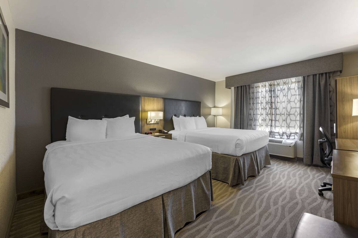 A hotel room with two beds, a desk, a chair, a lamp, and a window with curtains.