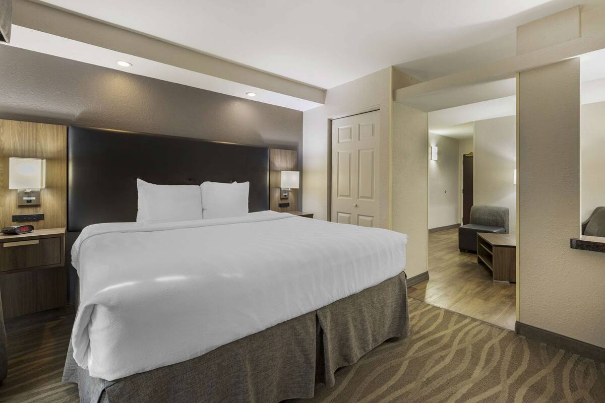 A hotel room with a large bed, bedside tables with lamps, a door, a seating area with a chair, and a mirror on the wall.