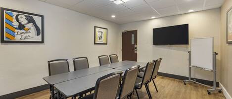 Uma sala de conferências com uma mesa comprida, cadeiras, um quadro branco e uma televisão na parede.