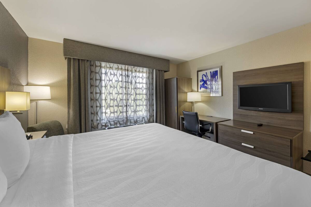 A hotel room with a large bed, a TV, a desk with a chair, and a window with curtains.
