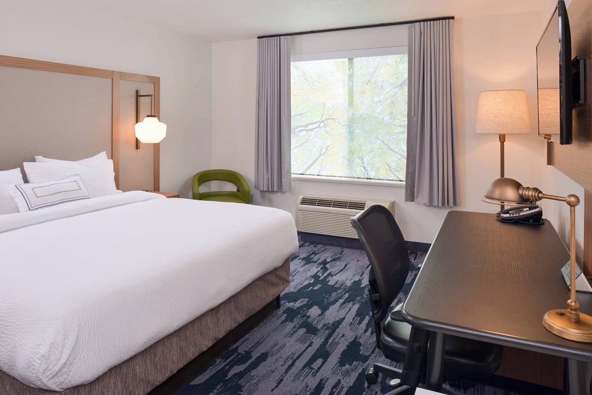 Une chambre d’hôtel avec un lit, un bureau, une chaise, une lampe, une télévision et une fenêtre avec des rideaux.