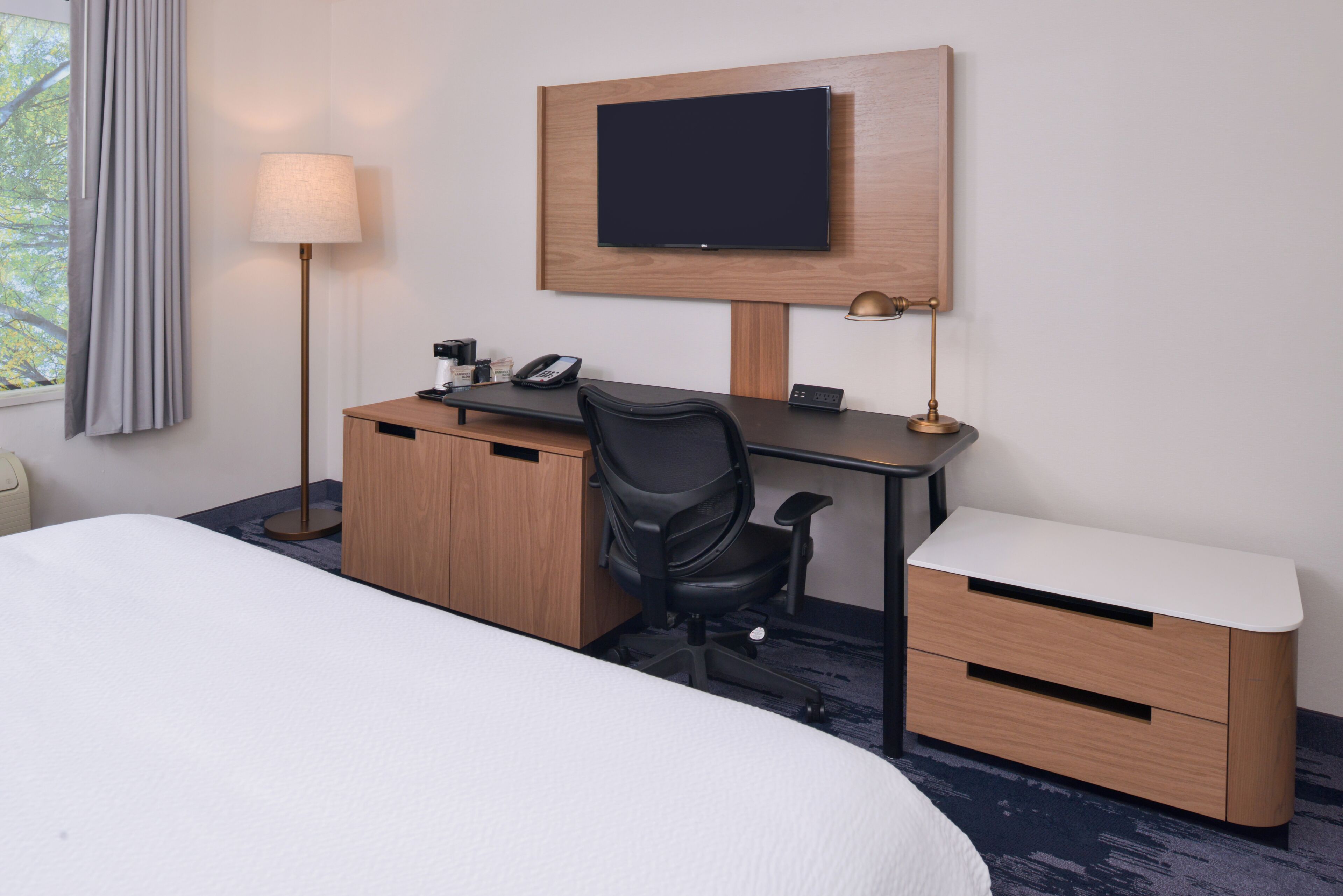 A hotel room with a bed, a desk with a chair, a TV mounted on the wall, a lamp, and a bedside table.