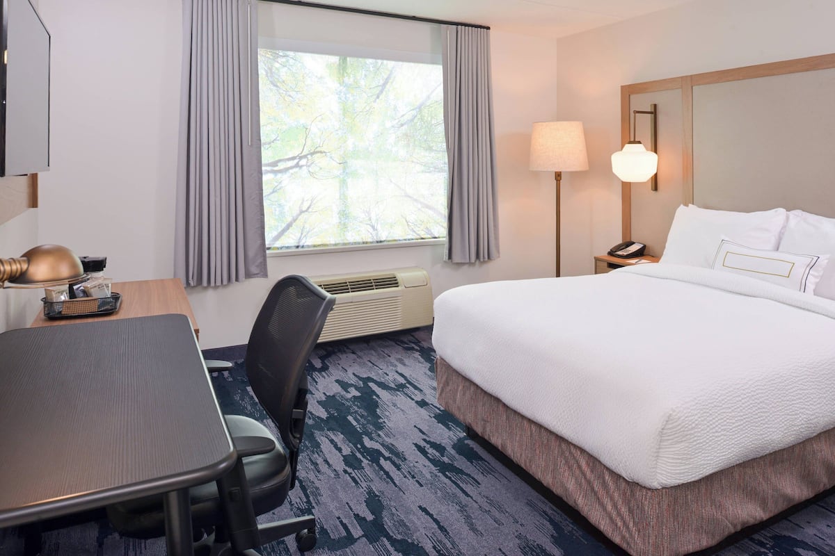 Une chambre d’hôtel avec un lit, un bureau, une chaise, une lampe et une fenêtre avec des rideaux.
