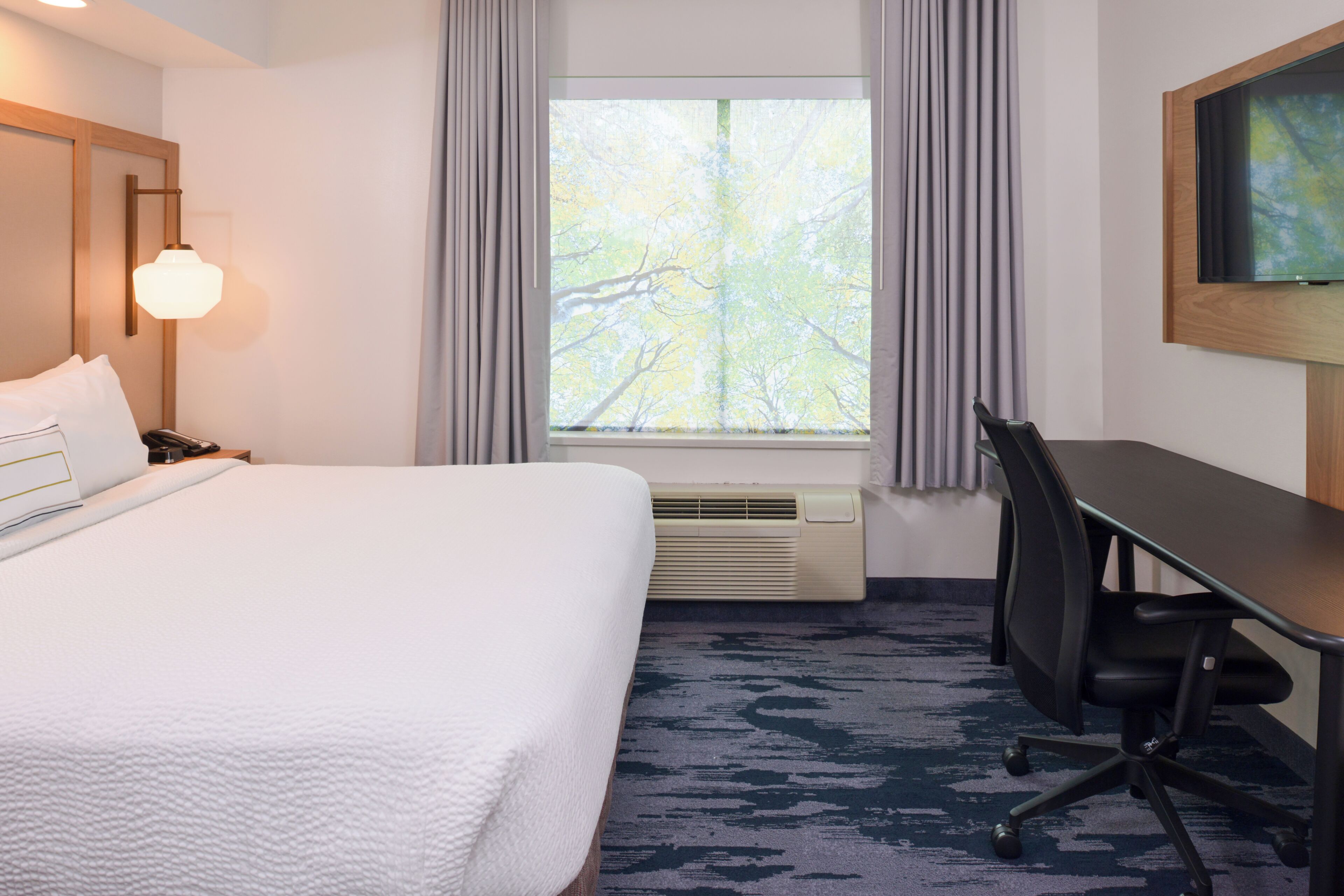 A hotel room with a bed, desk, air conditioning unit, and a window with curtains.