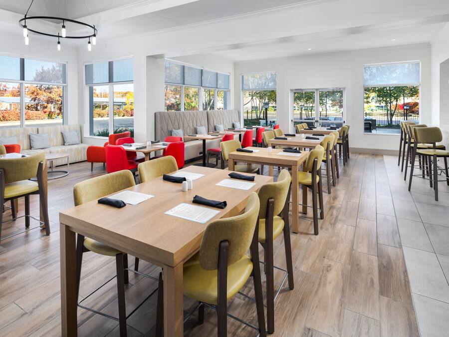 A modern dining area with multiple tables and chairs, large windows with a view of trees and a parking area.
