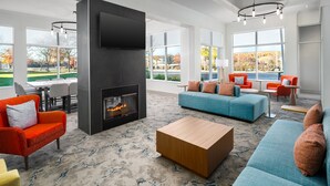 A modern living room with a fireplace, a large sofa, a coffee table, and a dining area with a view of the outdoors. - Hilton Garden Inn Rockaway (Rockaway)