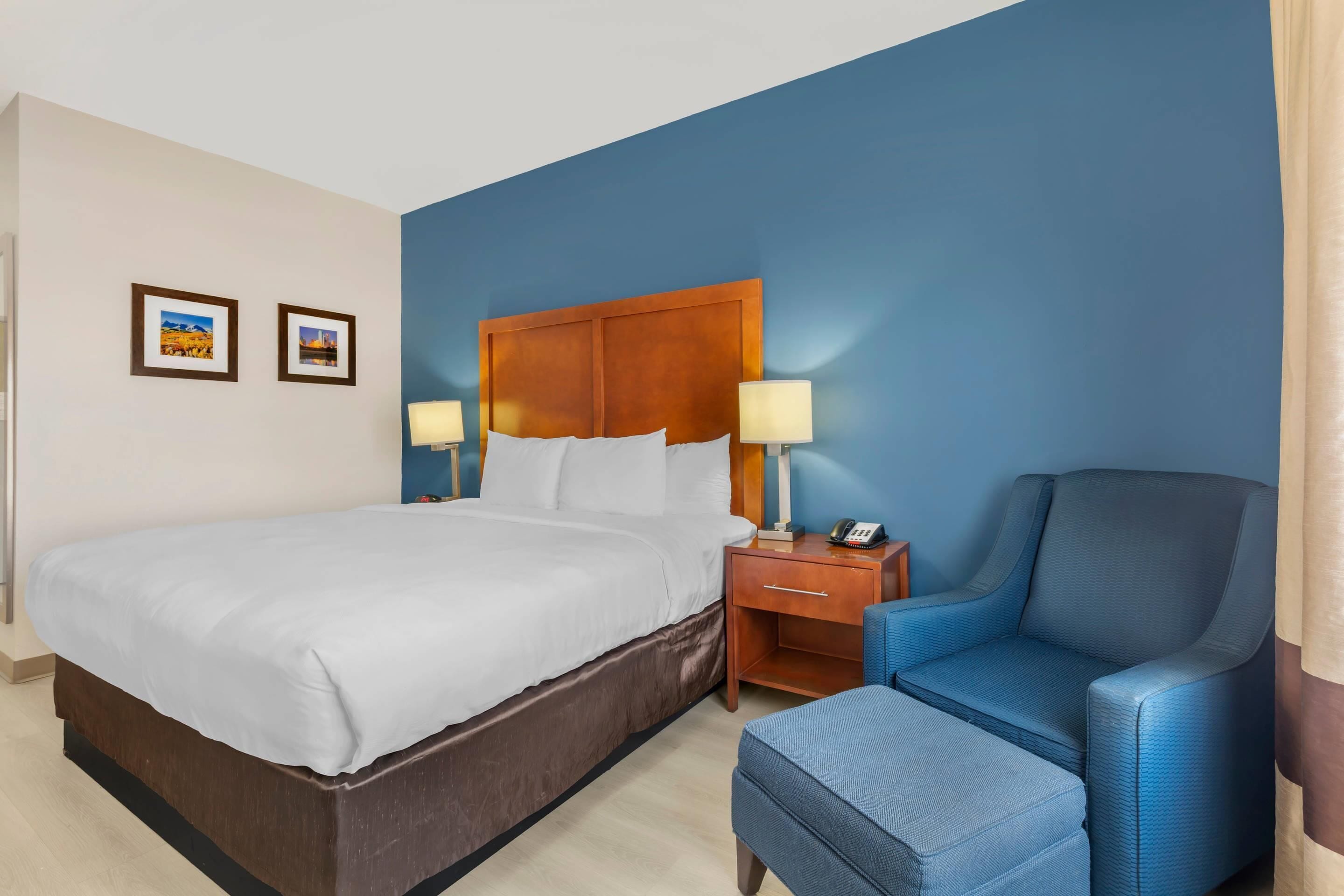 A hotel room with a large bed, two bedside tables with lamps, a blue armchair, and a small ottoman.