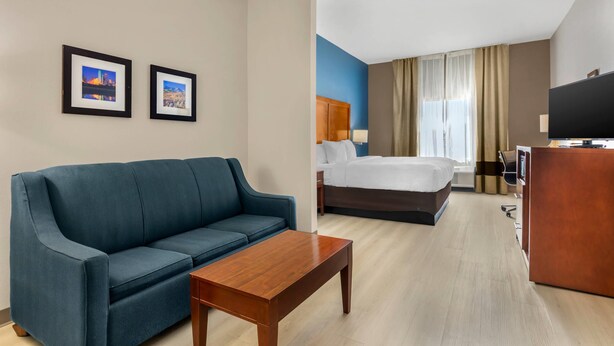 Une chambre d’hôtel avec un canapé bleu, une table basse en bois, un lit avec une literie blanche, un bureau avec une télévision et deux tableaux encadrés au mur.