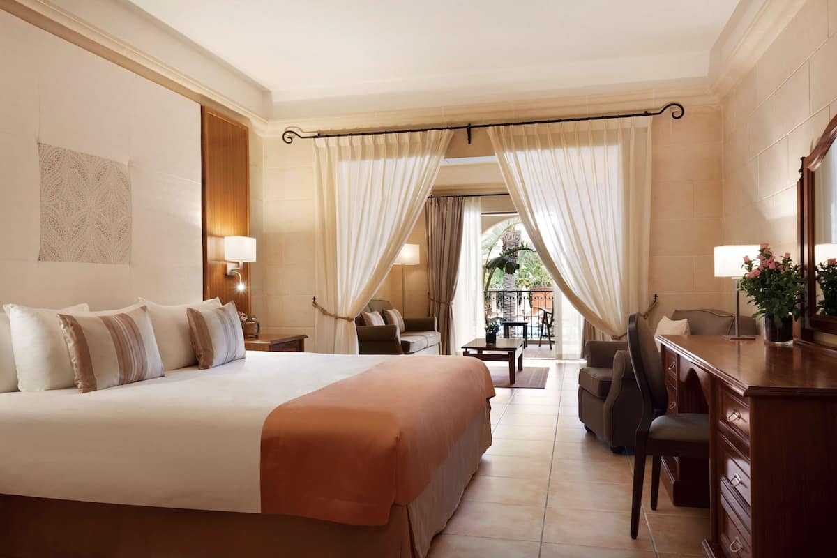 Ein Hotelzimmer mit einem großen Bett, einem Schreibtisch, einem Sessel und einer Lampe, mit Blick ins Freie.