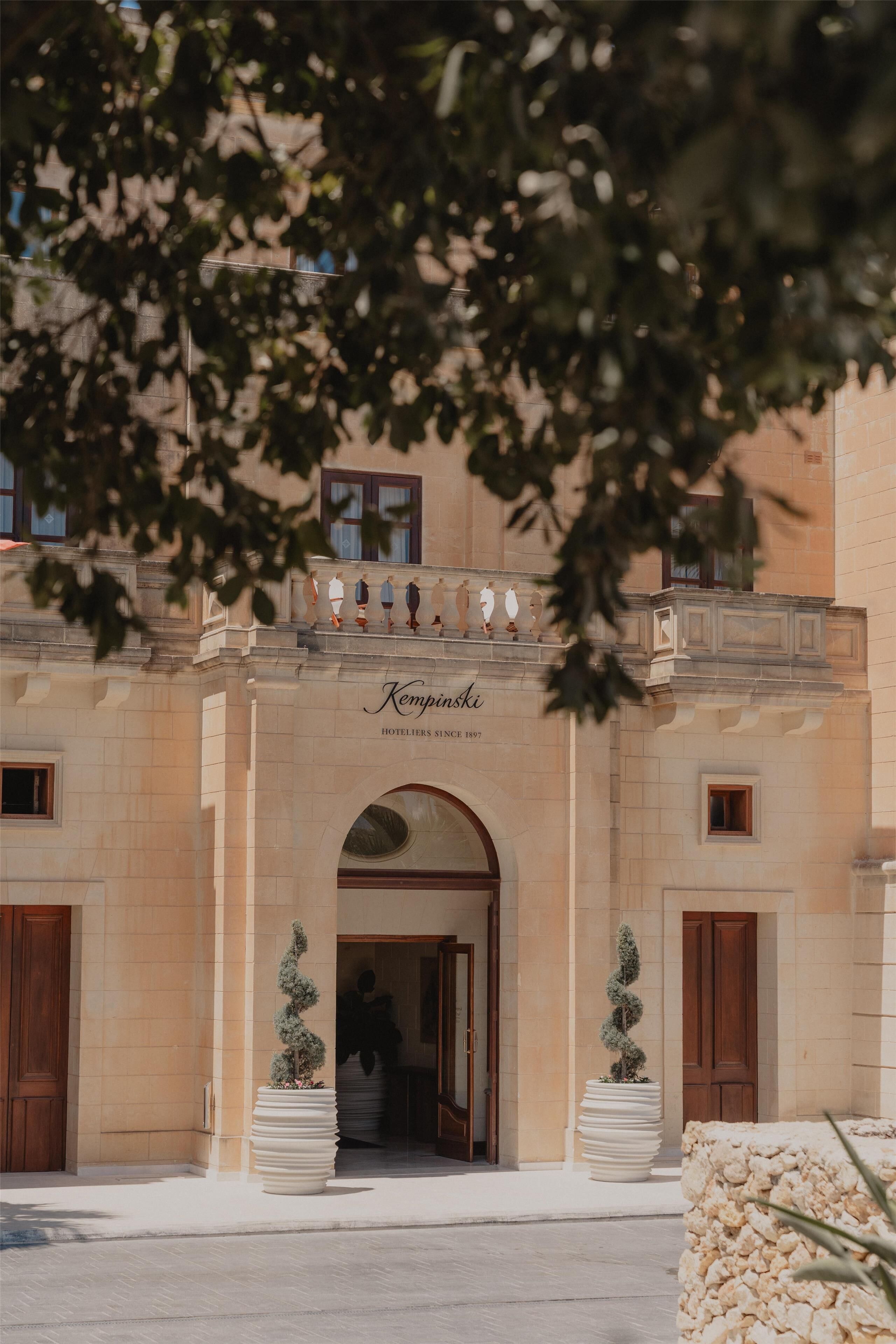 Entrada de hotel con fachada de piedra, un arco de entrada y dos macetas con plantas.