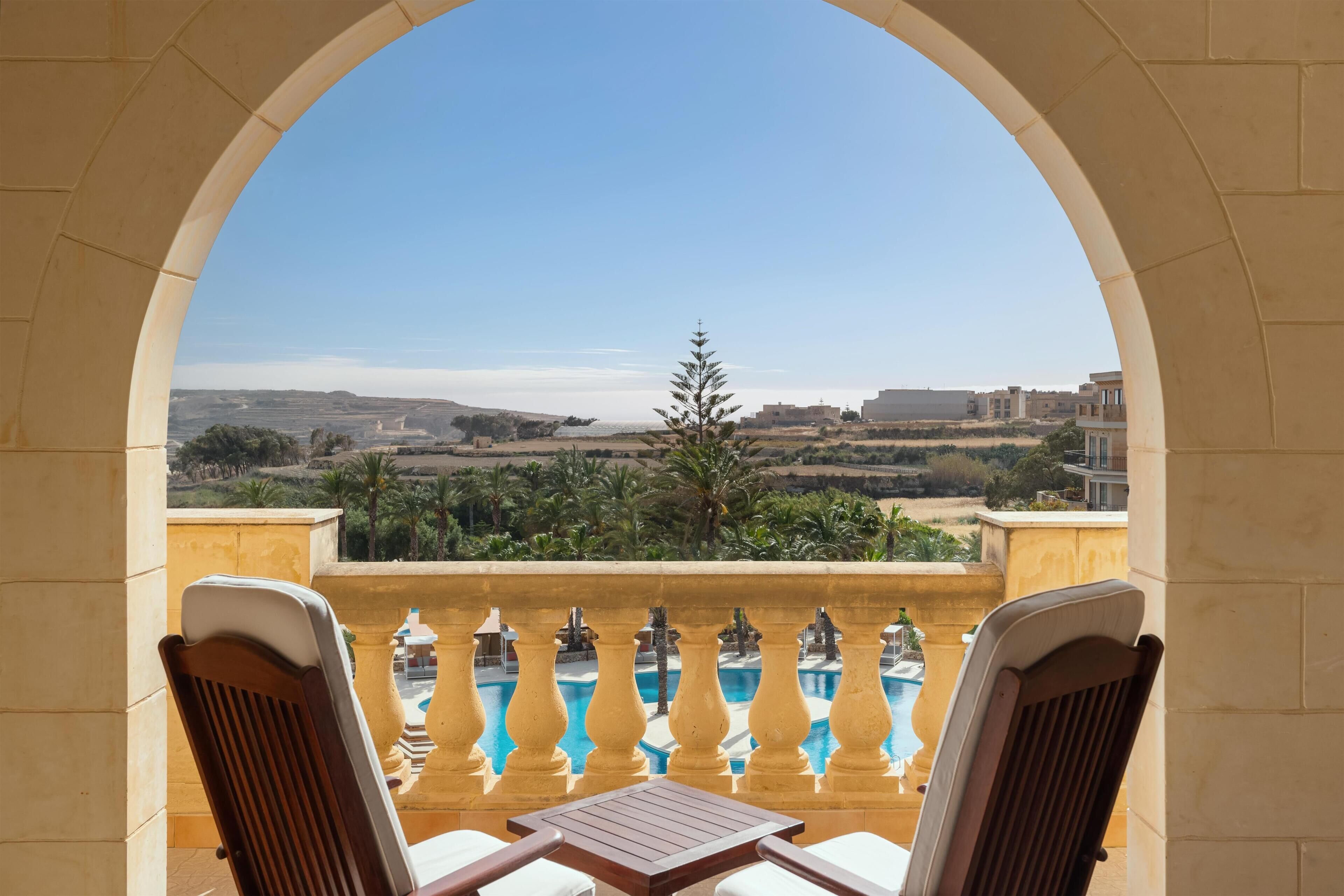 Un balcón con dos sillas y una mesa, con vistas a una piscina y un paisaje con árboles y edificios.