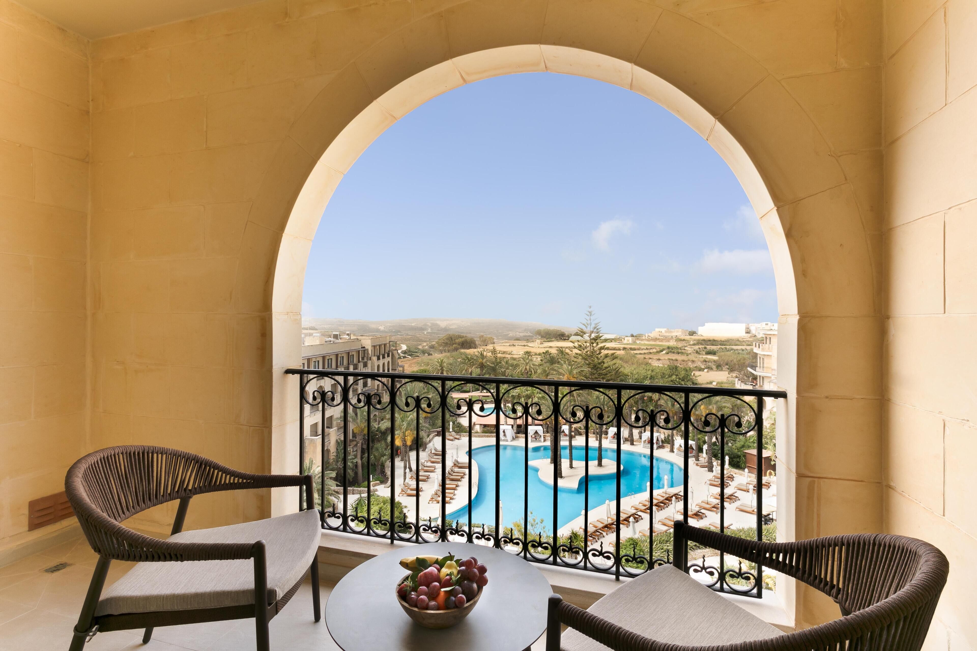 Un balcone con vista su una piscina, due poltrone in vimini, un tavolo con una ciotola di frutta e un parapetto in ferro battuto.