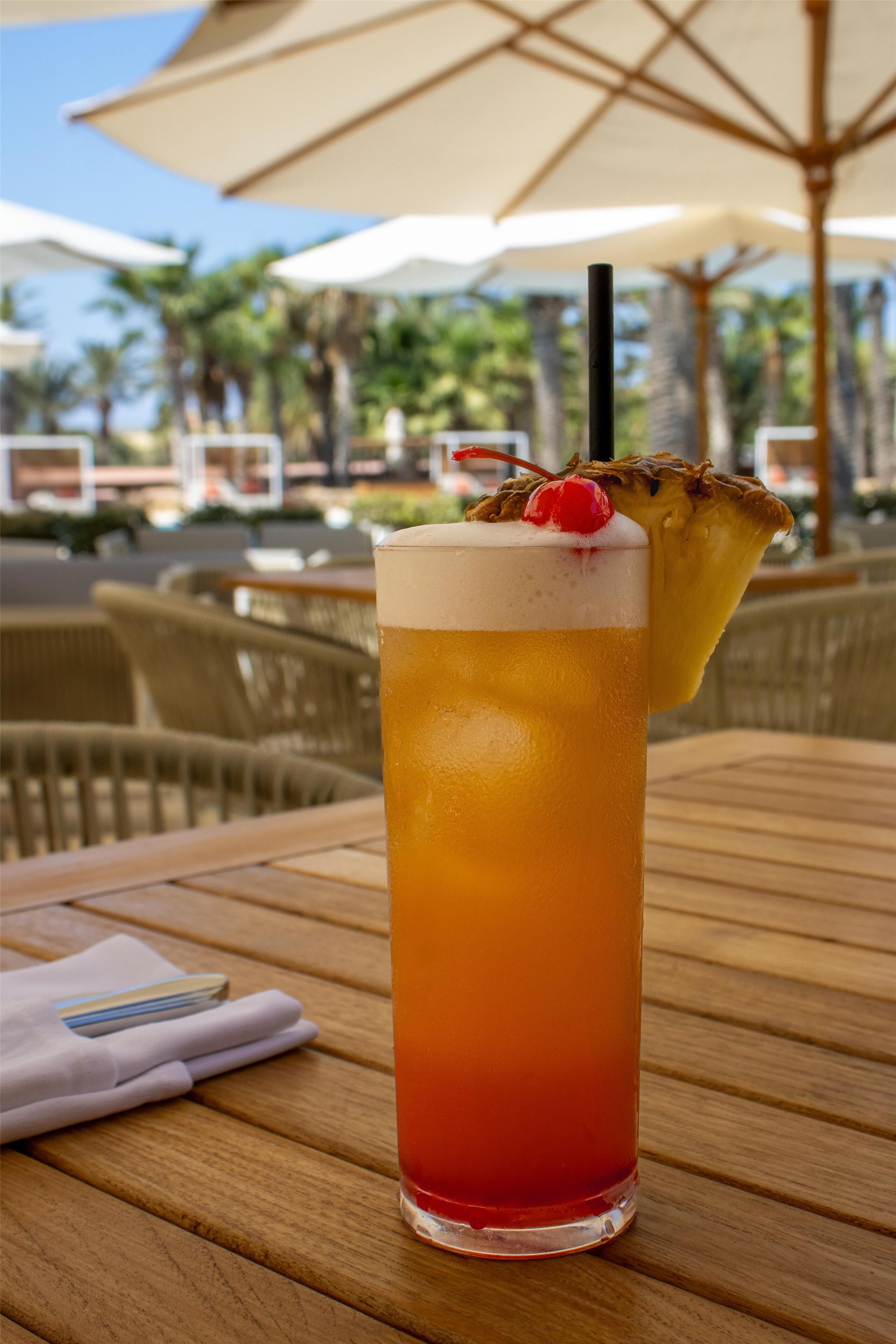 Un vaso alto con una bebida tropical, una cereza y una rodaja de piña en el borde, sobre una mesa de madera.