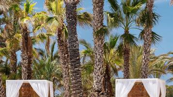 Un área junto a la piscina con dos cabañas blancas cubiertas, una piscina azul cristalina y altas palmeras.