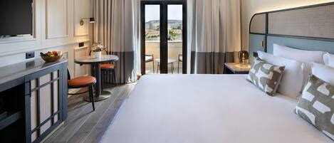 Habitación de hotel con cama, televisor, escritorio con silla, pequeña mesa con silla y balcón con cortinas.