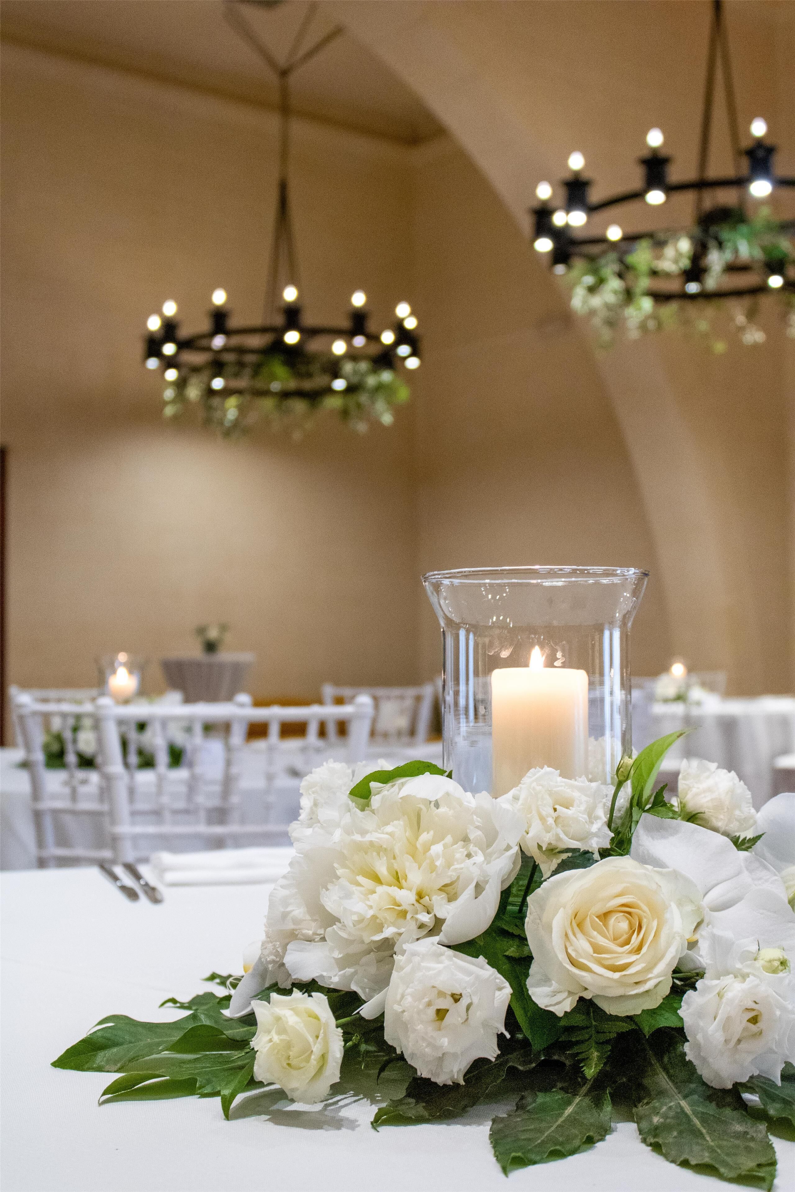 A table with white flowers and a candle in a glass holder, set in a room with chandeliers and white furniture.