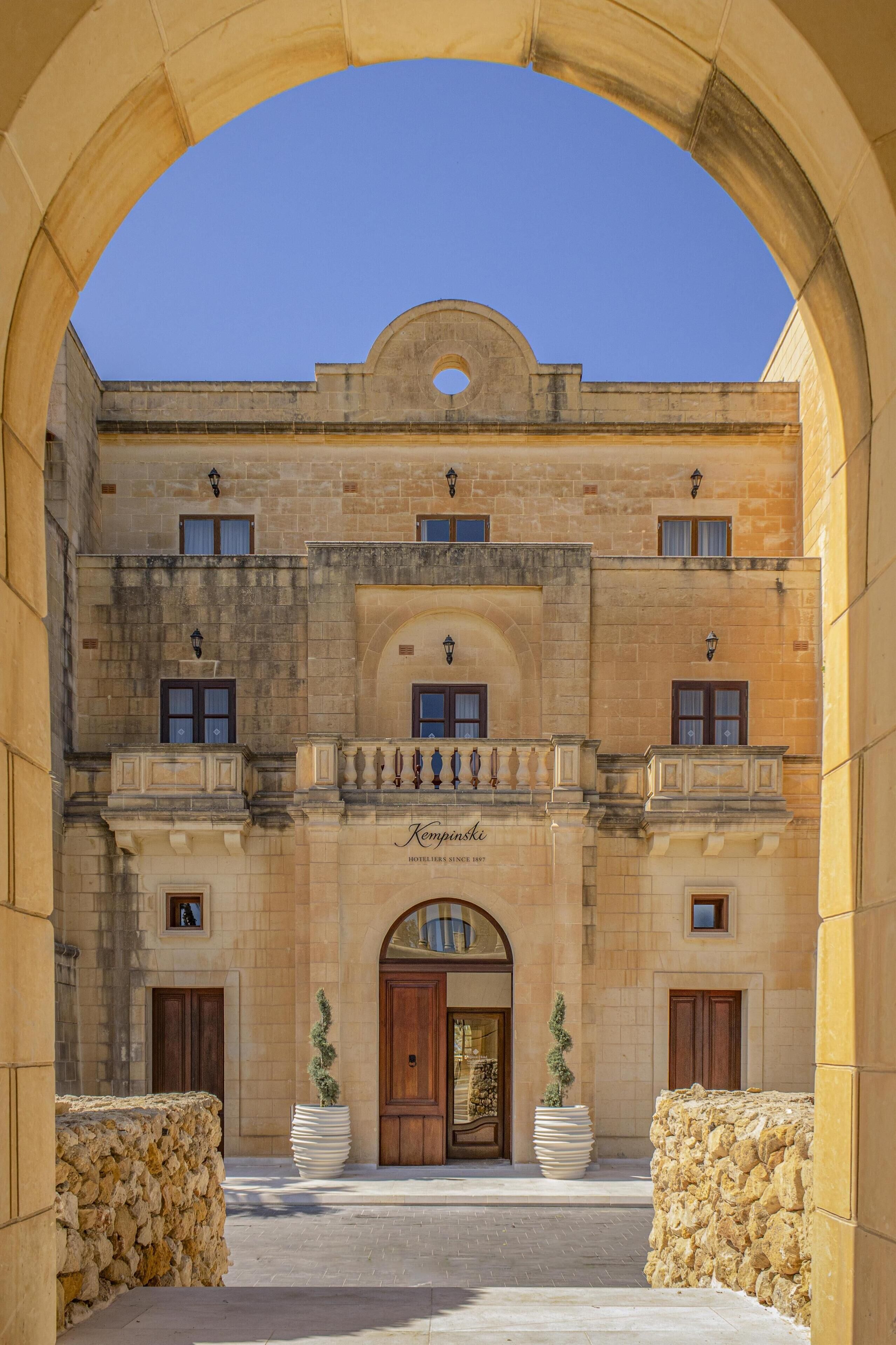 A historic building with a central arched entrance, stone walls, and a clear blue sky above.