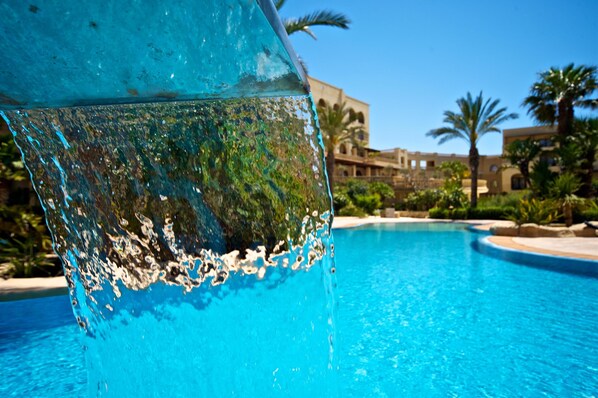 Vista despejada de una piscina con agua fluyendo, rodeada de palmeras y un edificio con arcos al fondo.