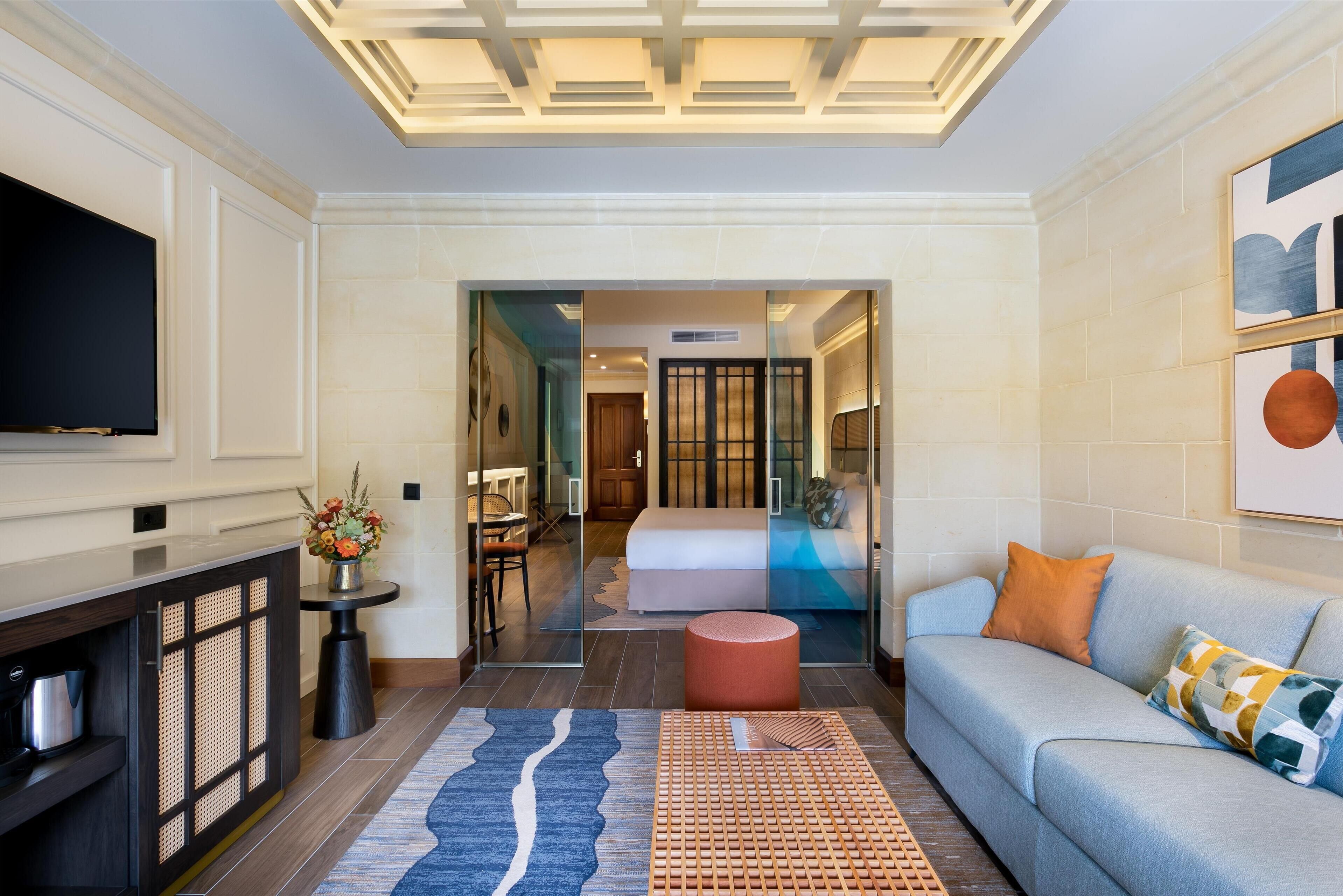 A modern hotel room with a flat-screen TV, a sofa, a coffee table, a decorative rug, and a glass partition leading to a bedroom with a bed and a bathroom.