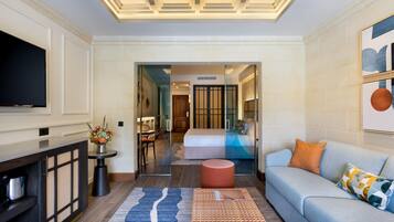 A modern hotel room with a flat-screen TV, a sofa, a coffee table, a decorative rug, and a glass partition leading to a bedroom with a bed and a bathroom.