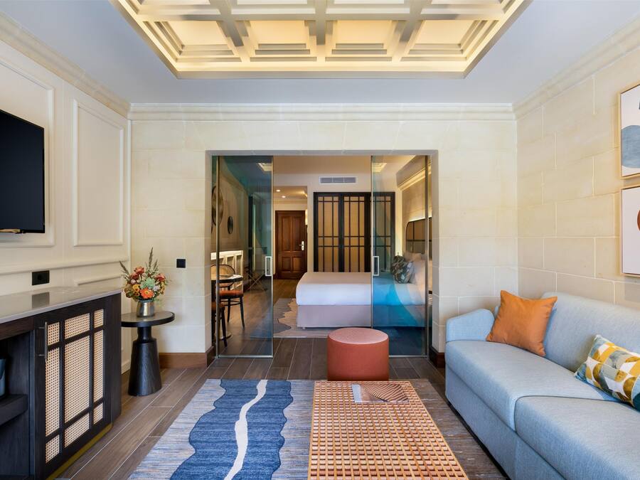 A modern hotel room with a flat-screen TV, a sofa, a coffee table, a decorative rug, and a glass partition leading to a bedroom with a bed and a bathroom.