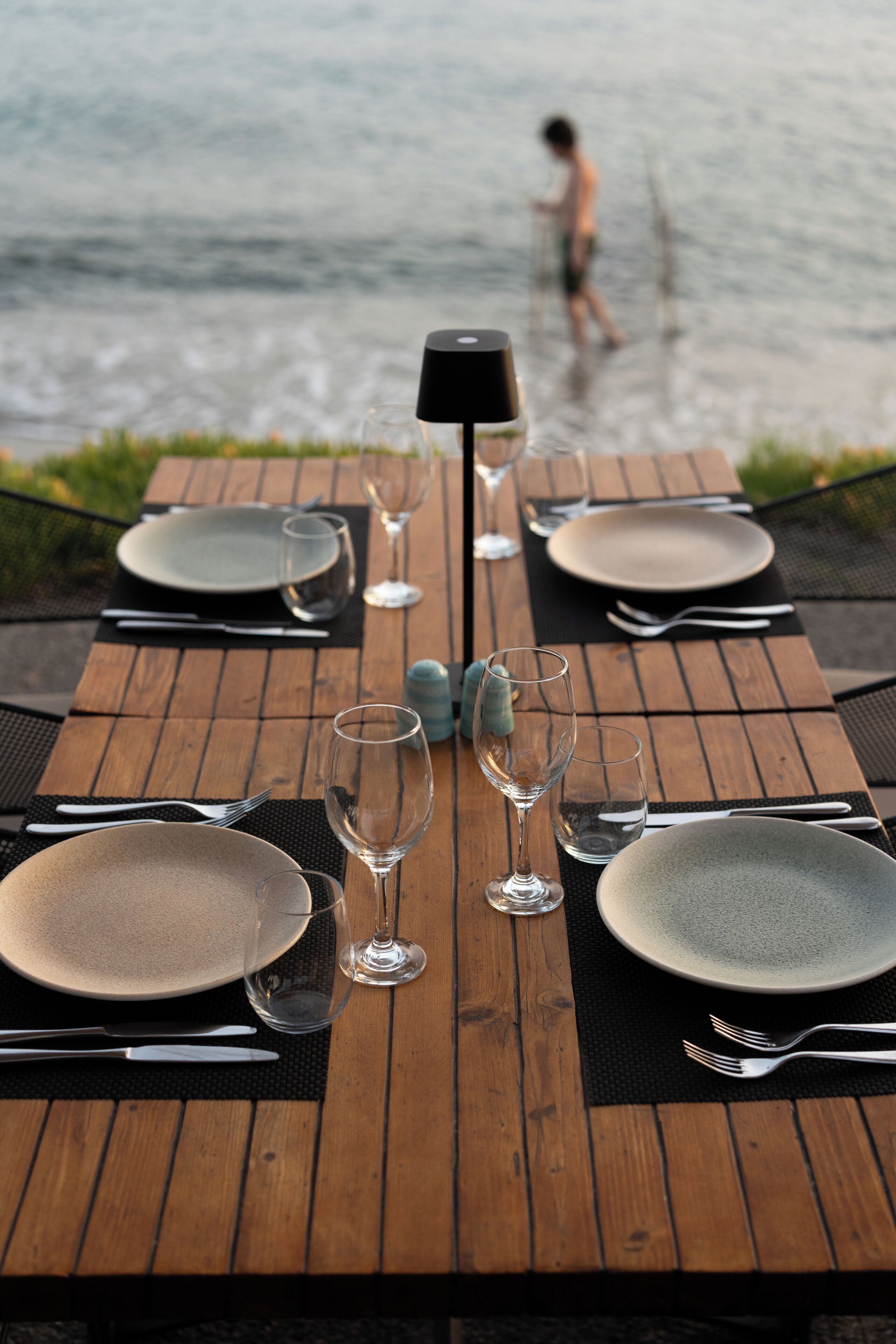 A wooden dining table set with plates, cutlery, and glasses, overlooking a body of water.