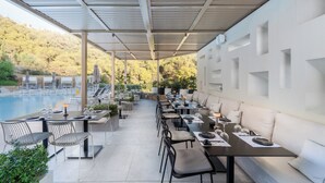 Un espace extérieur abrité, aménagé pour les repas, avec des tables et des chaises, une piscine en arrière-plan et une vue sur une colline.