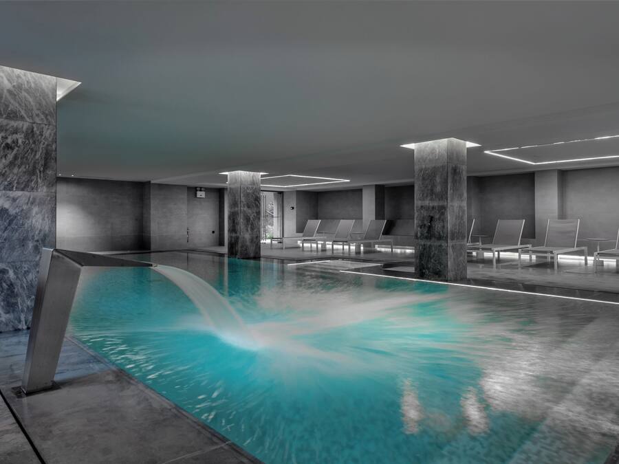 Una piscina interior moderna con columnas de mármol, sillones de descanso y una fuente de agua cristalina.