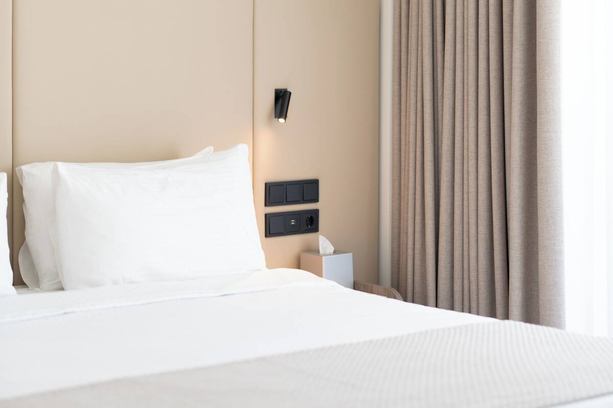 Une chambre d’hôtel avec un grand lit, une tête de lit dotée d’un éclairage intégré, une table de chevet avec une lampe et une fenêtre avec des rideaux.