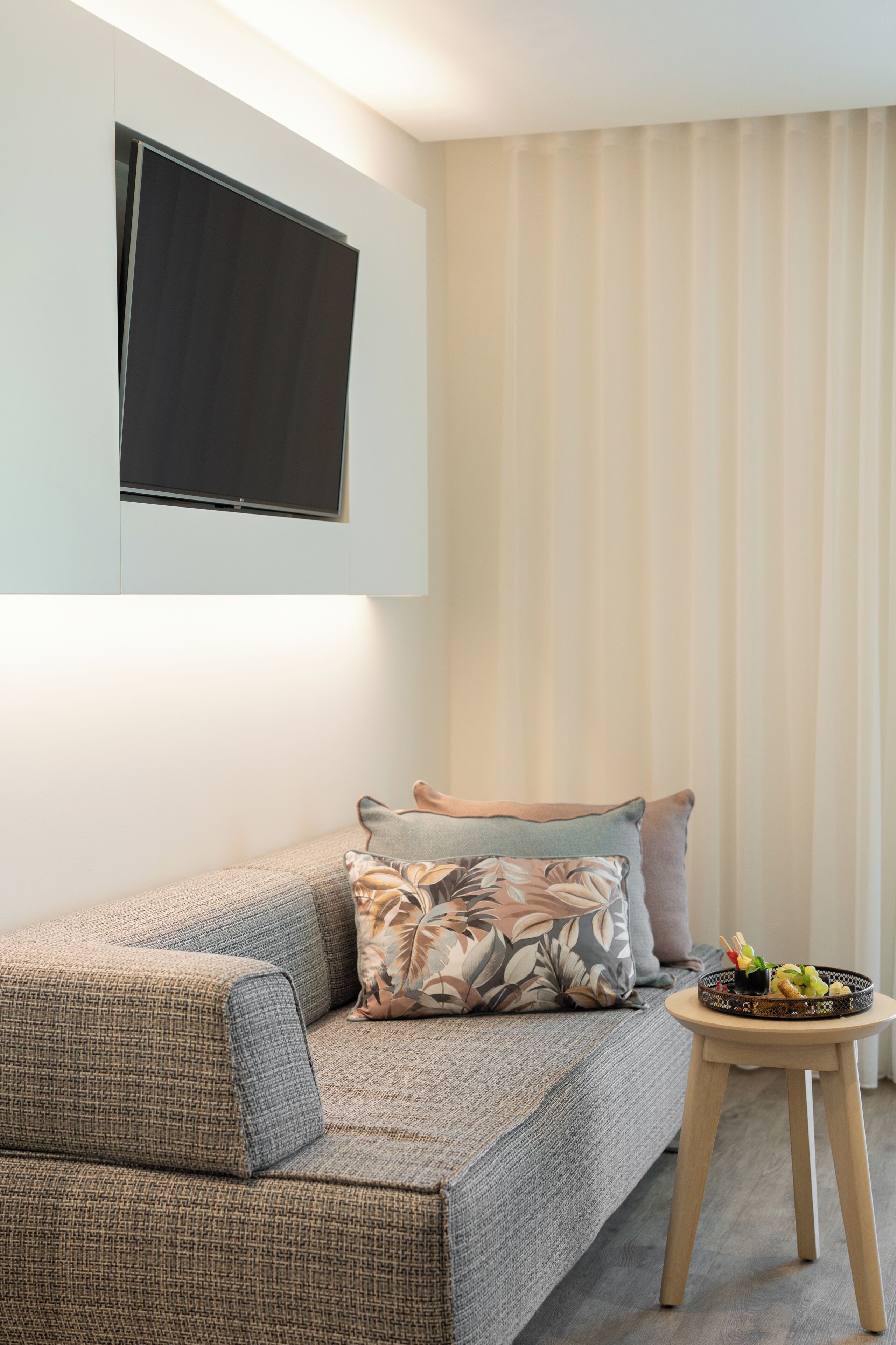 Een moderne woonkamer met een bank, een flatscreen-tv aan de wand, een klein houten bijzettafeltje met een fruitschaal, en een raam met verticale jaloezieën.