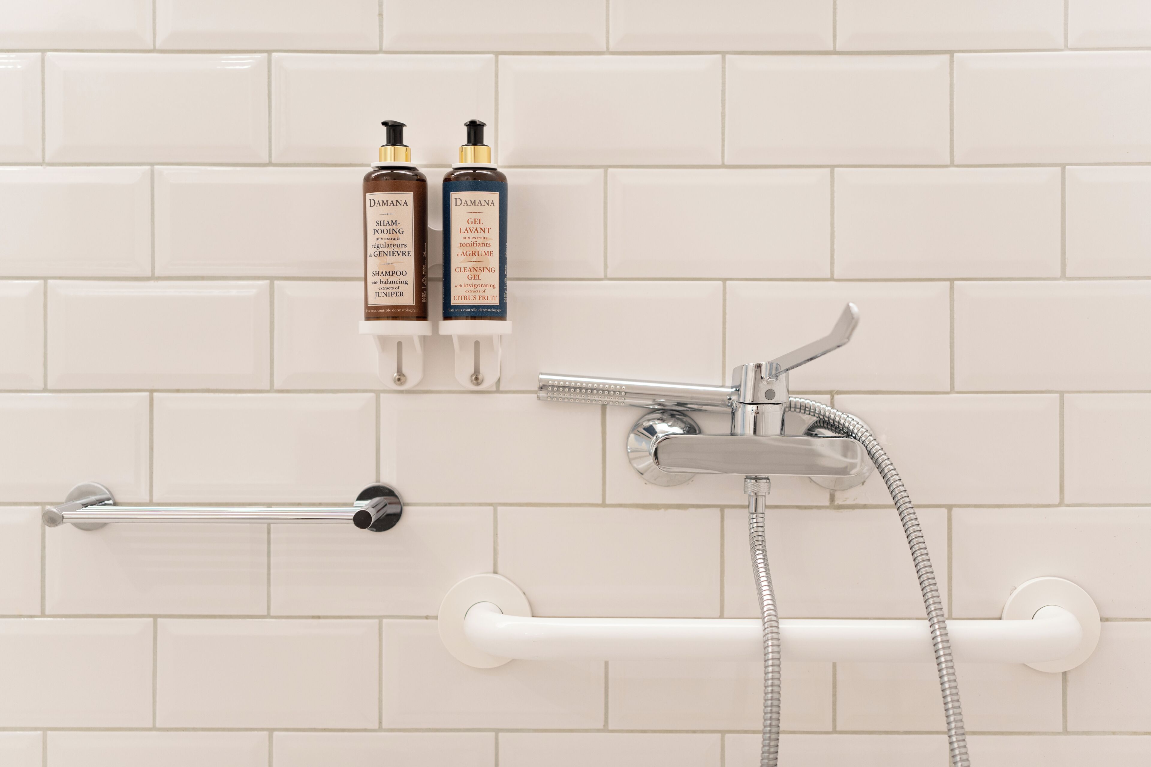 A bathroom with white tiles, a showerhead, and two bottles of shampoo and gel on the wall.