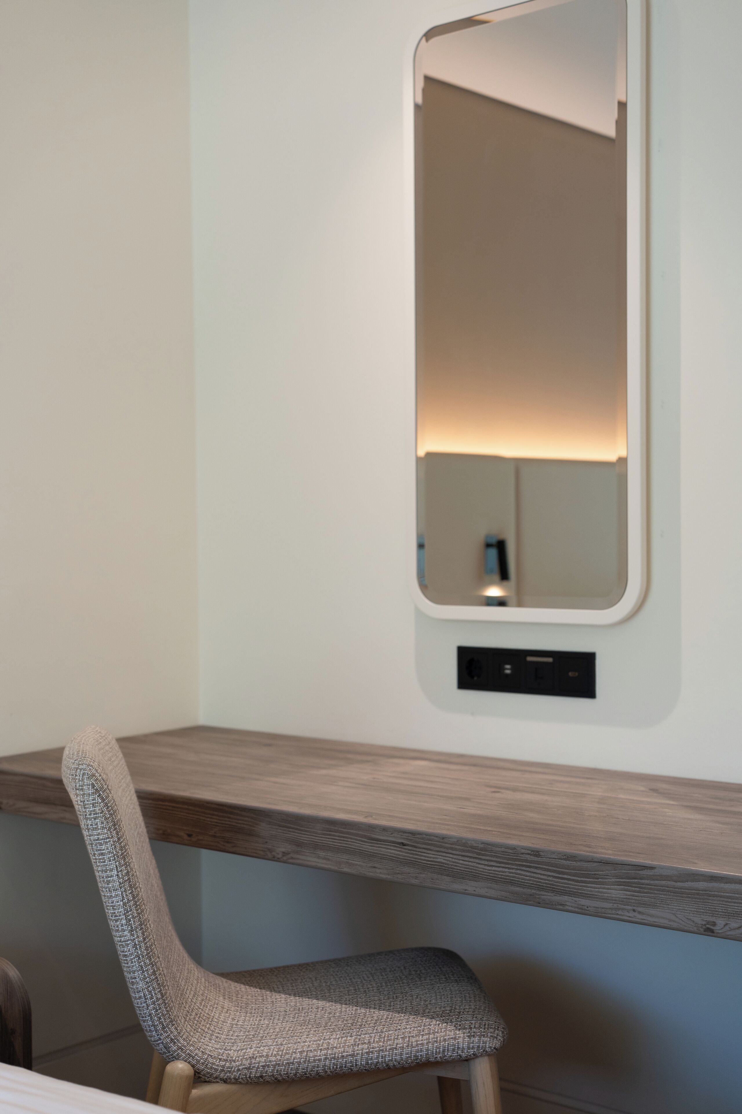 Una silla con asiento y respaldo tejido, un escritorio de madera con espejo incorporado, y un enchufe en la pared.