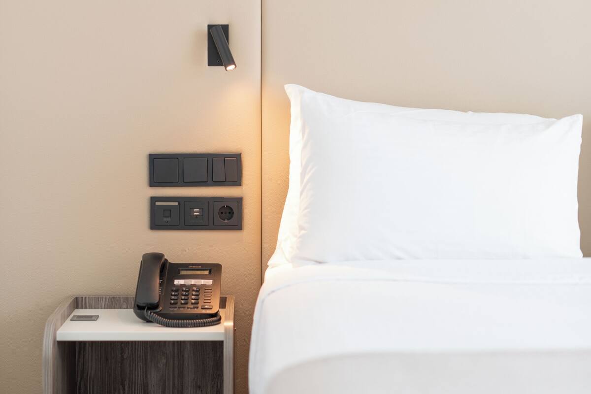 Une chambre d’hôtel équipée d’un lit, d’une table de chevet avec un téléphone, d’une lampe fixée au mur et d’une prise électrique.