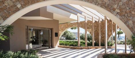 A stone archway leading to a building entrance with a glass door, surrounded by greenery and a tiled patio area.