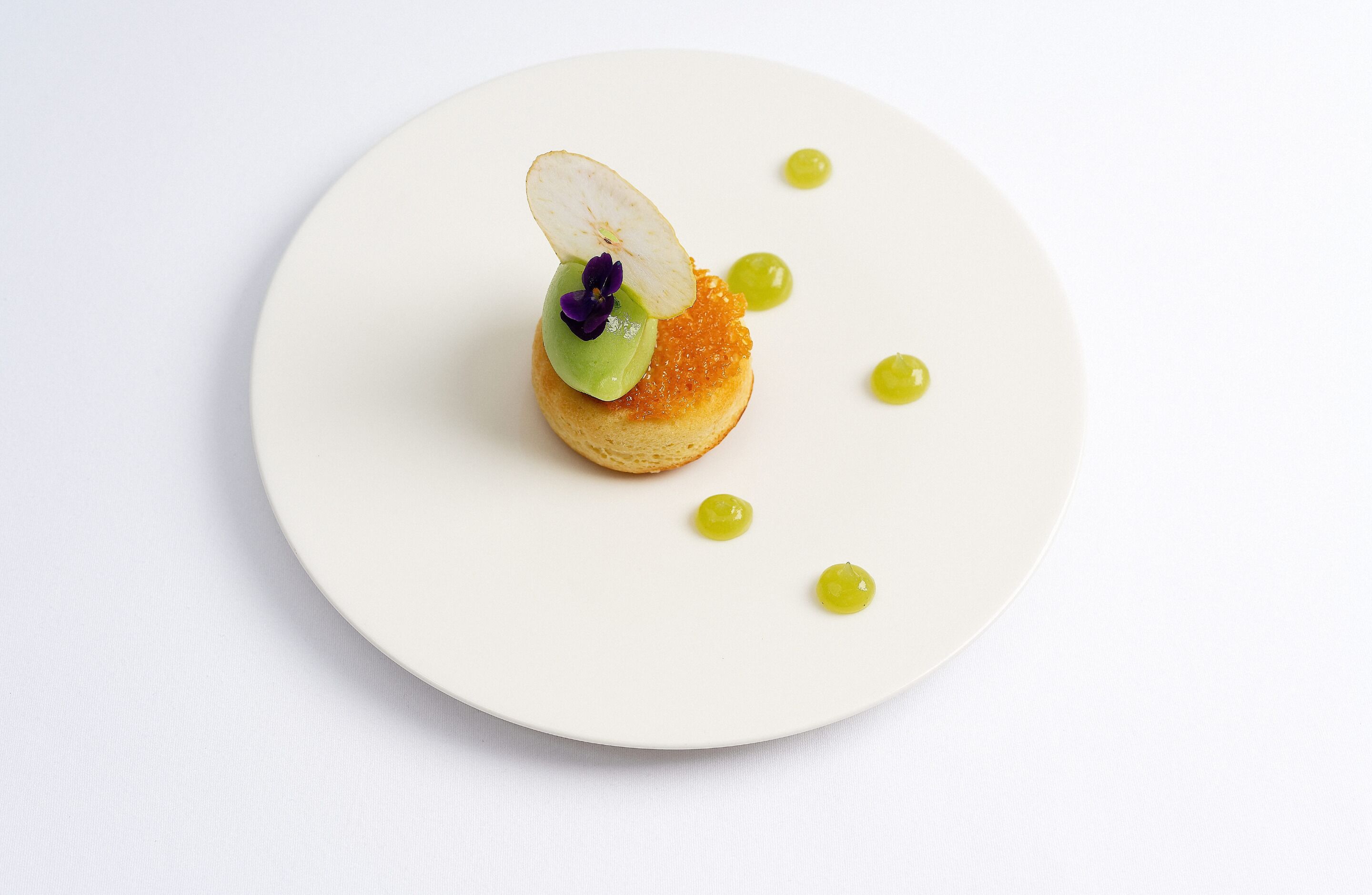Un plato blanco con un postre que incluye una base redonda pequeña cubierta con una porción de fruta, una cucharada de crema verde y unas escamas de caviar.